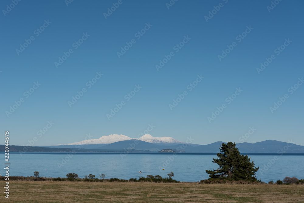 View across Lake Taupo, New Zealand, from the northern shore to the snow covered, volcanic peaks of Mounts Ruapehu, Tongariro and Ngauruhoe, under a clear blue, sunny sky.