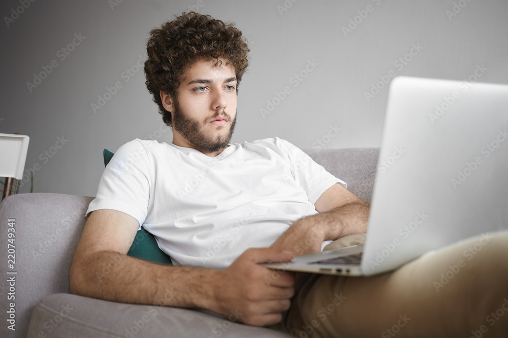 Image of confident young Caucasian journalist in casual wear having focused look while working remotely on portable computer, making research, lying comfortably on couch with gadget on his lap