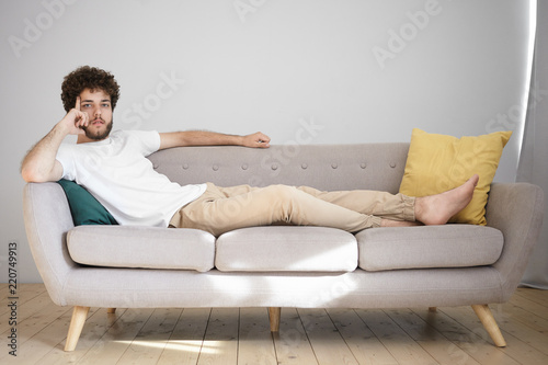 Attractive young Caucasian man with wavy hairdo and thick beard resting at home after work, lying barefooted on couch in his bachelor apartment, having pensive thoughtful expression, looking at camera