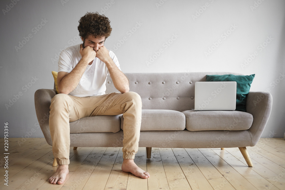 Horizontal shot of unemployed young male wearing white t-shirt and beige jeans sitting barefooted on sofa work portable computer having sad frustrated facial expression, searching for job online Horizontal shot of unemployed young male wearing white t-shirt and beige jeans sitting barefooted on sofa work portable computer having sad frustrated facial expression, searching for job online