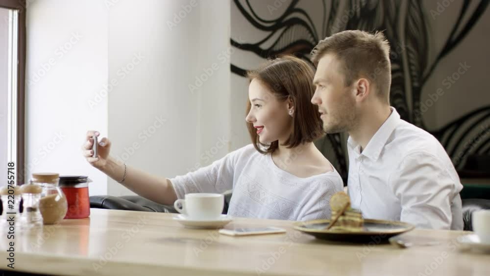 Two young people is sitting in a cafe. Man and woman are smiling and sitting in the cafe at the table during break. Couple is talking and drinking coffee. They are making selfies Two young people is sitting in a cafe. Man and woman are smiling and sitting in the cafe at the table during break. Couple is talking and drinking coffee. They are making selfies