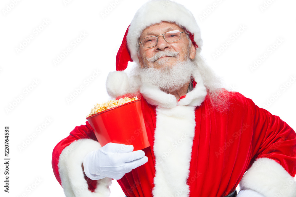 Naklejka premium Christmas. Smiling, kind Santa Claus in white gloves holds a red bucket with popcorn with one hand, and puts the other hand on his belt. The concept of visiting the cinema, watching a movie with