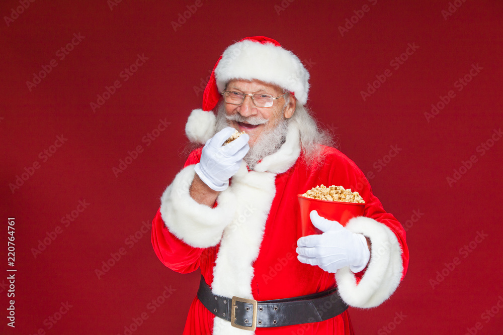 Christmas. Smiling, kind Santa Claus in white gloves with his mouth open holds a red bucket with popcorn with one hand, takes some popcorn and prepares to eat it with the second hand. The concept of