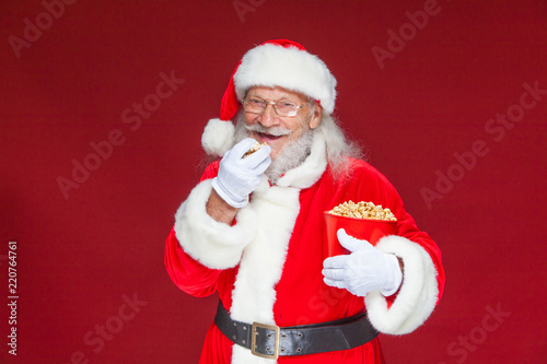 Christmas. Smiling, kind Santa Claus in white gloves with his mouth open holds a red bucket with popcorn with one hand, takes some popcorn and prepares to eat it with the second hand. The concept of