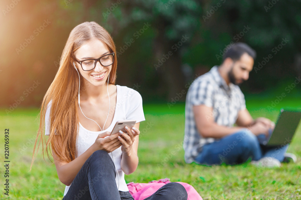 Attractive girl in white t-shirt wearing glasses sitting on the grass and writting message, her friend sitting at the back with a laptop