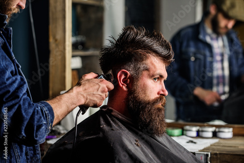 Barber styling hair of brutal bearded client with clipper. Barber with hair clipper works on hairstyle for bearded man barbershop background. Hipster lifestyle concept. Hipster client getting haircut