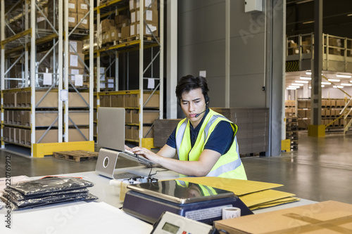 Confident young male customer service representative using laptop while sitting at desk in warehouse photo