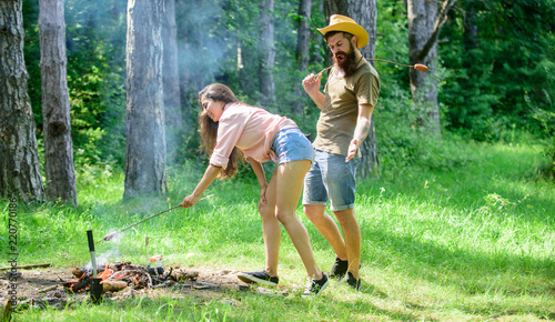 Man bearded hipster looks at sexy female buttocks. Girl bent down to fire while man enjoying sexy view. Couple picnic. Seductive position. Girl sexy buttocks denim shorts roasting sausage at bonfire