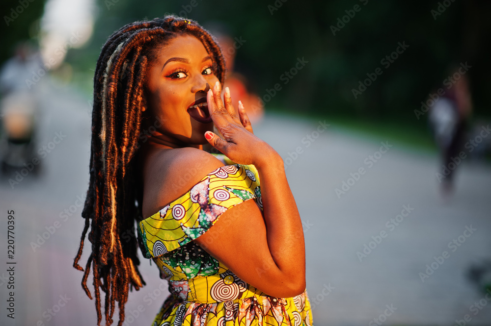 Cute small height african american girl with dreadlocks, wear at coloured yellow dress, posed at sunset, emotion surprise.