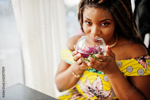 Cute small height african american girl with dreadlocks, wear at coloured yellow dress, sitting at cafe with flower at glass.