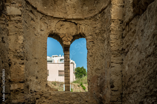 Inside of ancient stone brick ruin building with arched window and blue sky and buildings outside. © Pebo
