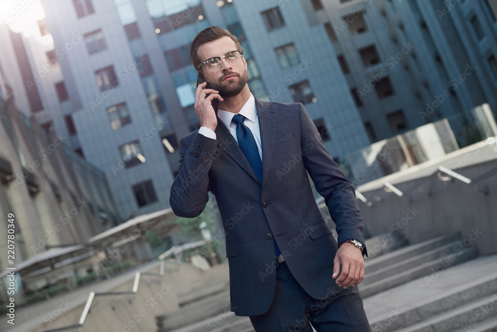 Business talk. Handsome young man in full suit talking on the phone and smiling while moving down the stairs outdoors Business talk. Handsome young man in full suit talking on the phone and smiling while moving down the stairs outdoors