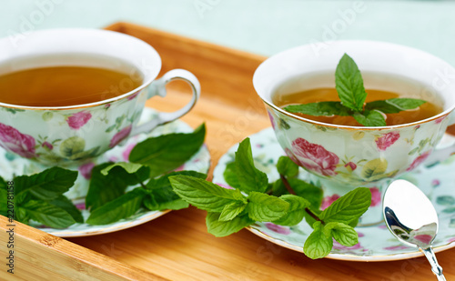 Mint herbal tea served in teacups with sacers and silver spoon with fresh leaves all around on wooden tray, copy space