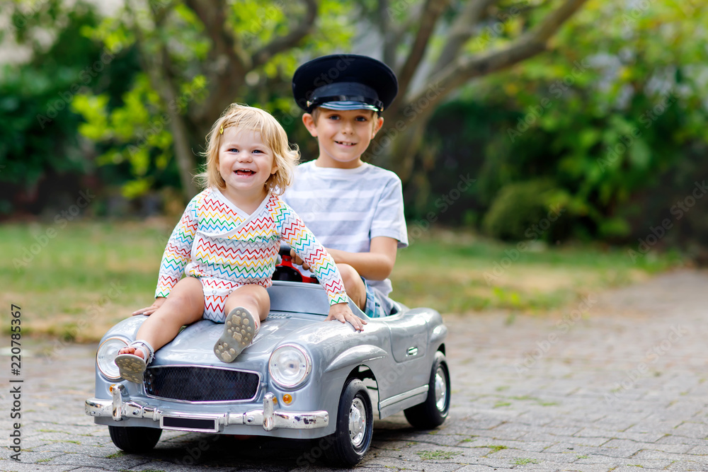 Two happy children playing with big old toy car in summer garden, outdoors. Kid boy driving car with little toddler girl inside. Laughing and smiling kids. Family, childhood, lifestyle concept Two happy children playing with big old toy car in summer garden, outdoors. Kid boy driving car with little toddler girl inside. Laughing and smiling kids. Family, childhood, lifestyle concept