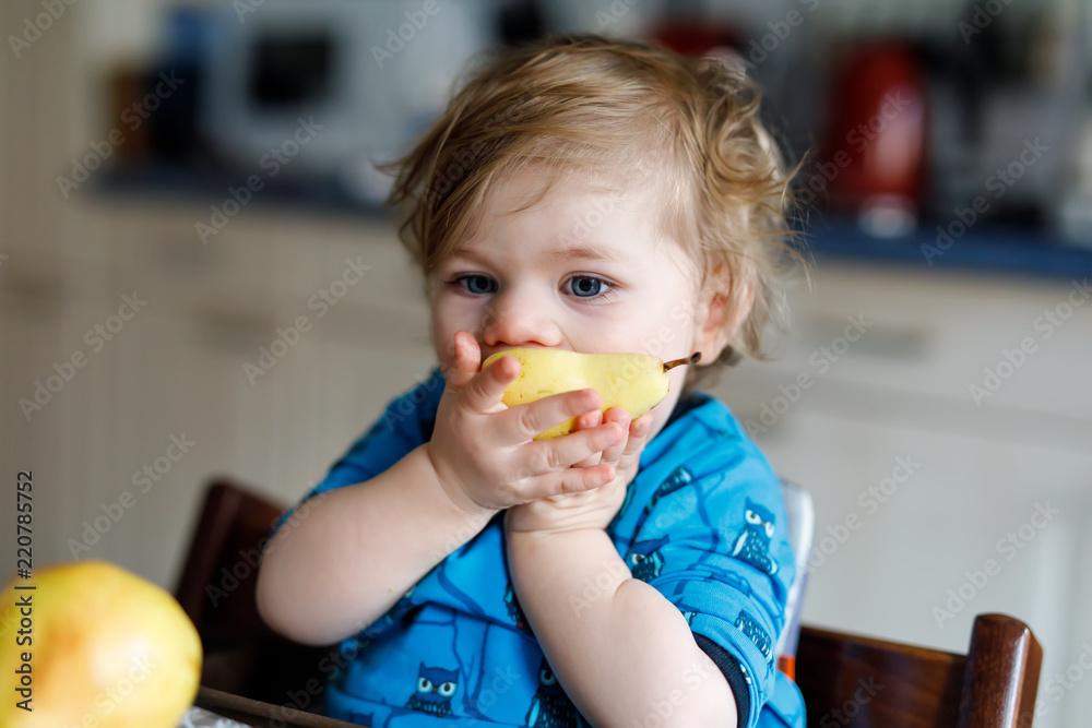 Cute adorable toddler girl eating fresh pear . Hungry happy baby child of one year holding fruit. Girl in domestic kitchen, having healthy meal snack. Smiling blond kid