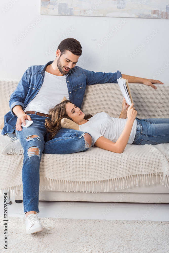 smiling man with smartphone looking at girlfriend while she reading book on couch at home