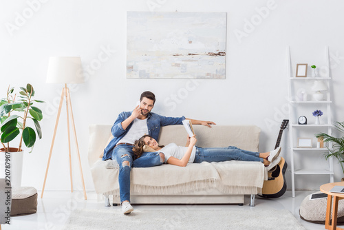 young man talking on smartphone while his girlfriend reading book on sofa at home