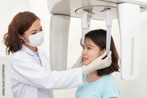 Asian woman in mask and uniform making panoramic dental x-ray of young smiling girl in office