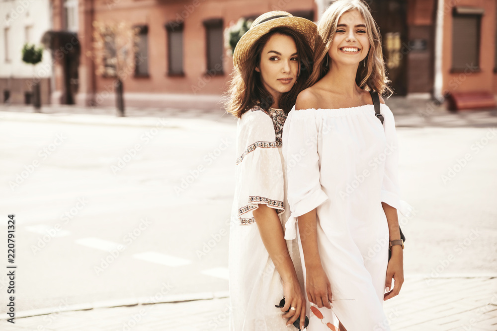 Naklejka premium Fashion portrait of two young stylish hippie brunette and blond women models in summer sunny day in white hipster clothes posing on the street background. No makeup