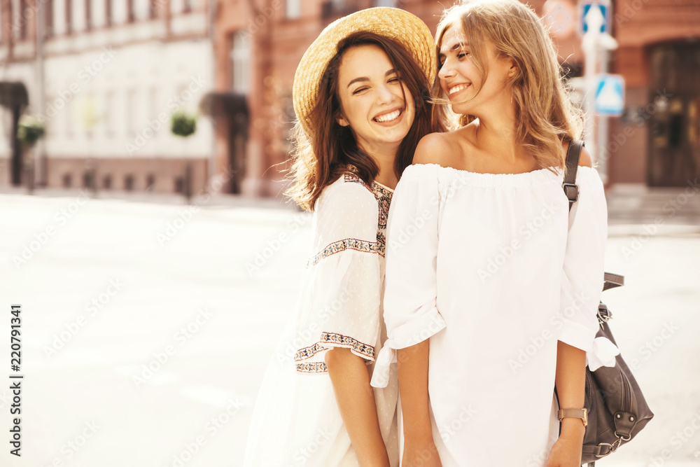 Obraz premium Fashion portrait of two young stylish hippie brunette and blond women models in summer sunny day in white hipster clothes posing on the street background. No makeup