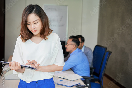 Beautiful Asian woman standing in office room with coworkers on background and using tablet 