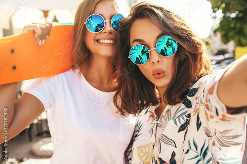 Two young female stylish hippie brunette and blond women models in summer hipster clothes taking selfie photos for social media on smartphone on the street background. With colorful penny skateboards photo