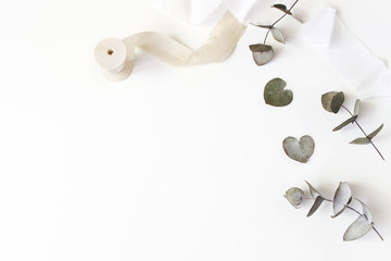 Feminine wedding desktop scene. Composition of dry silver dollar eucalyptus leaves and silk ribbons on white table background. Flat lay, top view.