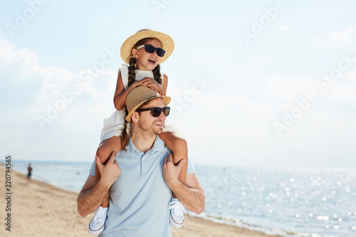 Waist up portrait of modern young man holding daughter on shoulders and walking on beach along sea enjoying Summer vacation, copy space