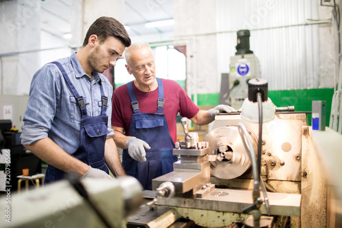 Young engineer looking at rotating detail of industrial machine while his senior colleague explaining how it works