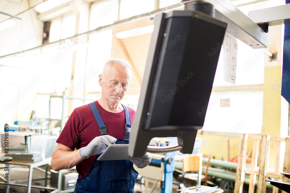 Aged engineer in overalls searching for online information in tablet while standing in front of monitor Aged engineer in overalls searching for online information in tablet while standing in front of monitor