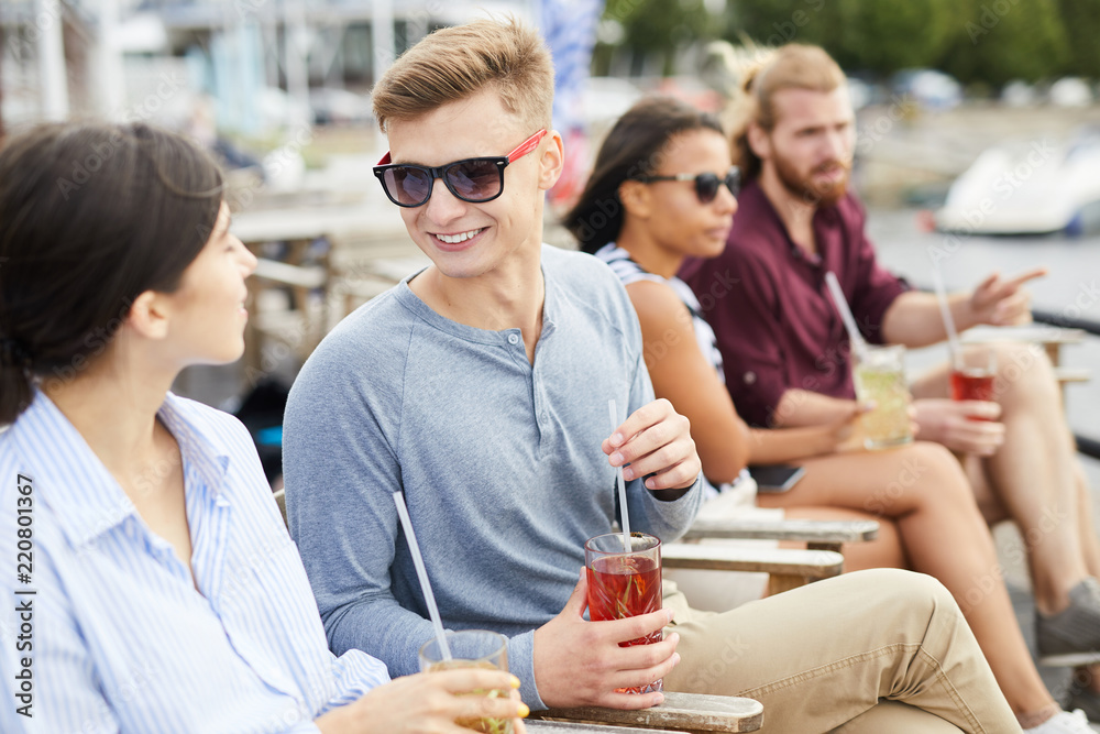 Smiling guy in sunglasses having drink while talking to his girlfriend on summer vacation