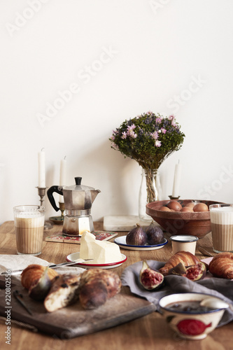 Breakfast in modern kitchen with croissants, figs, coffee, bread on board over rustic wooden table background, empty space on white wall