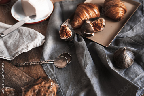 Breakfast with croissants, figs, spoons, batter, bread on wooden board over rustic wooden background, ceramics dishes, warm colors, above view photo