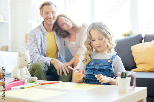 Cute little girl with curly hair drawing with highlighters with her parents on background