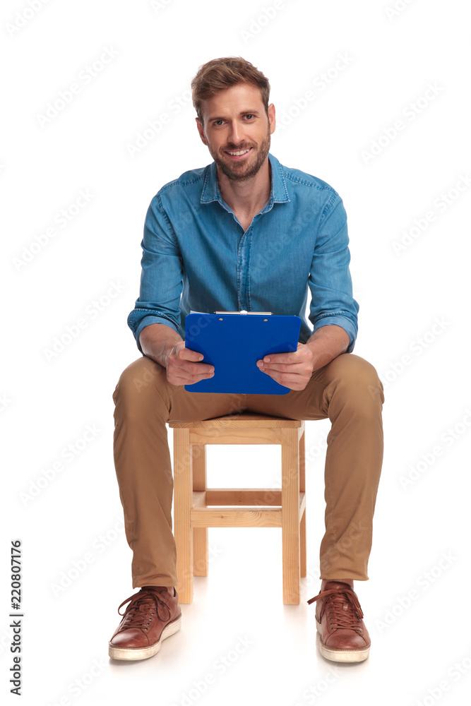 smiling student sits on wooden chair and holdis clipboard smiling student sits on wooden chair and holdis clipboard