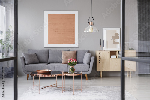 Flowers on copper table in front of grey couch in living room interior with rose gold poster. Real photo