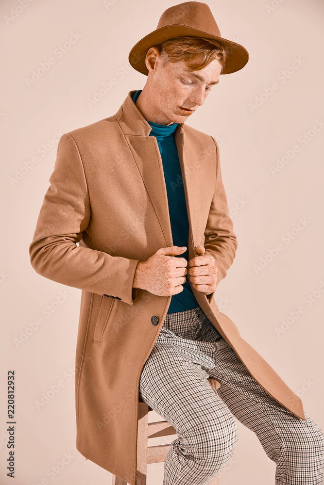 Young male model with red hair and freckles posing in brown coat and hat,  plaid pants, turquoise sweater, sitting on chair. Pink background Stock  Photo | Adobe Stock