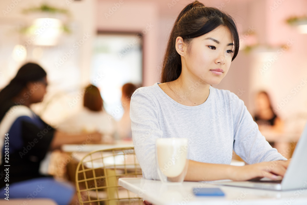 Asian girl typing on laptop keypad and looking at its display while browsing in cafe after college Asian girl typing on laptop keypad and looking at its display while browsing in cafe after college