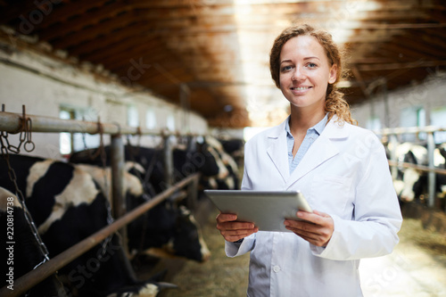 Waist up portrait of cheerful female veterinarian smiling looking at camera while using digital tablet standing in cowshed of modern dairy farm, copy space photo