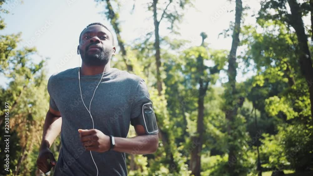 Happy african american man run listen music in headphones using the phone smile at park smartphone training active athlete body exercise fit health sport workout runner summer app muscular jogging