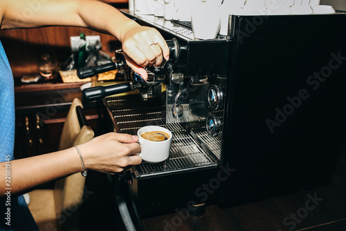 Barista is making black coffee americano. Girl is holding a white cup in her hands near the professional coffee machine. Food and cafe shop concept.