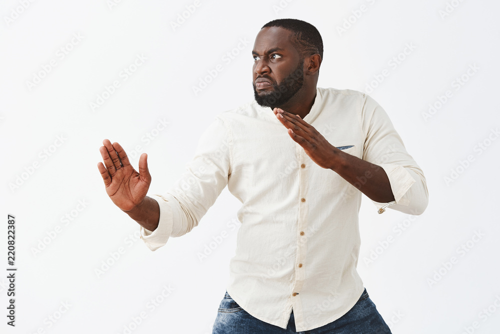 Funny and playful adult African American guy with beard, standing in profile in martial arts pose with raised palms, frowning and staring left with angry and evil expression to fight with enemy Funny and playful adult African American guy with beard, standing in profile in martial arts pose with raised palms, frowning and staring left with angry and evil expression to fight with enemy