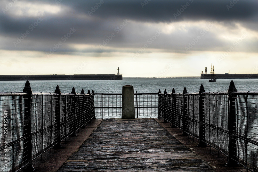 A slatted concrete pier overlooks the rough sea of Dover in England, on a gray day while the sky takes on shades of yellow and white