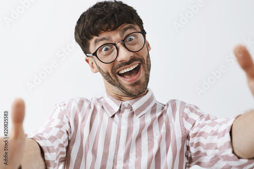 Close-up shot of funny emotive and happy good-looking modern male hipster in glasses and pink striped shirt pulling hands towards camera to make selfie, aping and making faces, fooling around