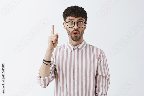 Waist-up shot of thrilled and excited attractive young hipster guy in glasses and pink shirt raising index finger in eureka finger folding lips, gasping adding important suggestion having great idea