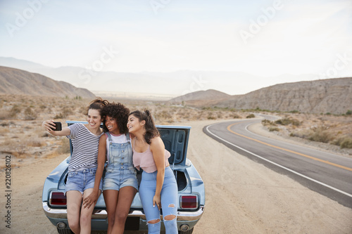 Three Female Friends Posing For Selfie Sitting In Trunk Of Classic Car On Road Trip