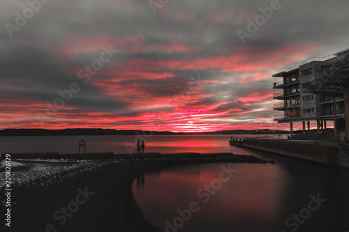 Norwegian sunset taken from the Astrup Fearnley museum in Oslo, the red colors of the sun go out behind clouds behind the sea, while the shadows of people playing on the beach are in motion photo