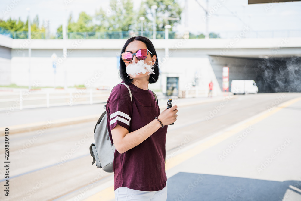 Obraz premium Vaping. Young beautiful woman smoking e-cigarette with smoke outdoors. Vapor concept.
