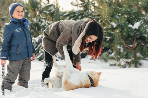 Little child and mom plays with funny Akita-inu dog in a winter park. Christmas happy family,mother and son walking with dog lying on snow in winter day. Drinking hot coffee or tea on snowy winter