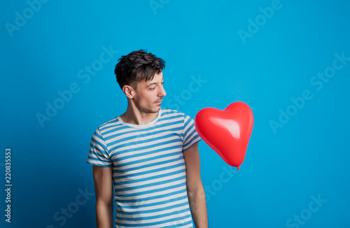 Portrait of a young man in a studio on a blue background, looking at red heart.